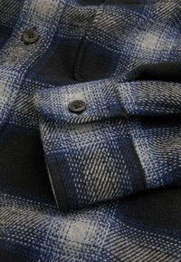 Blaut und graues Karostoff mit einer weichen Textur, zwei schwarzen Knöpfen und einem gefalteten Kragen mit einem karierten Muster.