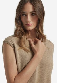 Stickad beige ärmlös topp med ribbad rund halsringning, texturerat tyg och subtila axelsömmar. Närbild av modellens hand på tyget.