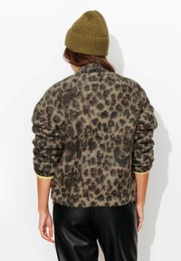 Pull-over en tissu duveteux à imprimé léopard dans des tons neutres avec poignets côtelés. Le modèle porte un bonnet vert olive et un pantalon noir. Tissu doux et texturé.