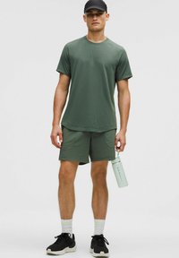 Uomo che indossa una maglietta sportiva verde oliva, pantaloni corti abbinati, cappellino nero e sneaker, mentre tiene in mano una bottiglia d'acqua verde chiaro.