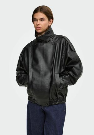 Jeune personne aux cheveux courts portant une veste noire oversize en simili-cuir avec un col montant et un jean bleu, mains dans les poches.