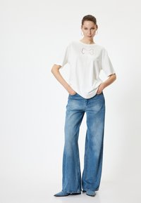 Witte korte mouwen t-shirt met geborduurde strikdetails, gecombineerd met hoge taille wijdvallende blauwe jeans. Model staat met handen in de zakken.