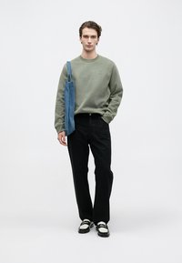 Mlad moški v zeleni kapuci in črnih hlačah, ki nosi modro torbo na rami, stoji pred belo ozadjem.