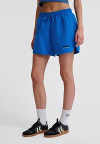 Blå idrottsshorts med dragsko i midjan, sidofickor och svart "hummel" logotyp. Bärs med svarta sneakers och vita strumpor.