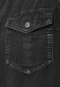 Mustast teksariidest kangas, millel on rinnatasku tipuga klapiga, metallist nupu detailid ja tekstuuriga õmblused. Värvilt peenelt tuhmistunud.