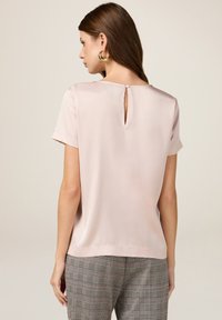 Top in satin rosa a maniche corte con scollatura rotonda e chiusura a occhio di bottone sul retro, abbinato a pantaloni a quadri. Texture liscia, design minimal.
