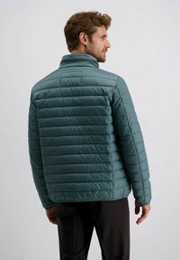 Man in een groene gewatteerde jas die zijwaarts staat, met de rug en profiel zichtbaar, in donkere broek tegen een effen lichte achtergrond.