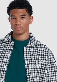 Camicia a quadri bianca e nera con colletto, dotata di taschino sul petto e bottoni bianchi, indossata sopra una maglietta color teal.
