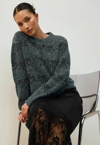 Szary puszysty sweter z strukturalnym wzorem i subtelnym połyskiem, w zestawieniu z czarną koronkową spódnicą. Modelka siedzi na przezroczystym krześle.