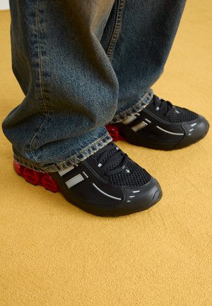 Black athletic shoes with a mesh upper, glossy accents, and red cushioning at the sole, paired with blue denim jeans.