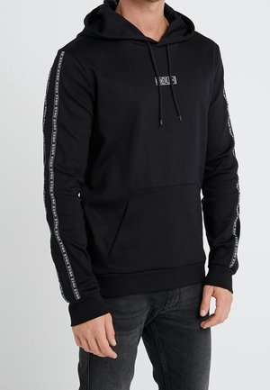 Hombre con sudadera negra con capucha y bolsillo delantero, texto blanco en el pecho y franjas de logotipo verticales en las mangas.