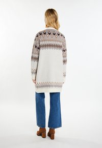 Langer, hellgrauer Cardigan mit geometrischen Mustern in Braun und Creme. Getragen über blauen, weit geschnittenen Jeans und braunen Stiefeletten mit Absatz.