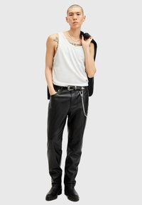 Zwarte leren broeken met een kettingdetail, gecombineerd met een wit tanktop. Het model houdt een zwart jasje vast en staat tegen een neutrale achtergrond.