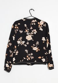 Czarna kurtka z długim rękawem w beżowe i białe kwiatowe wzory, wisząca na czarnym wieszaku na białym tle.