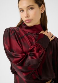 Blusa rossa in satin con colletto alto e maniche a sbuffo, caratterizzata da un motivo floreale e un orlo arricciato, che mette in risalto una ricca texture e design.