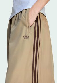 Béžové športové šortky vyrobené z ľahkej látky s tromi tmavohnedými pruhmi po bokoch a malým logom na prednej strane.