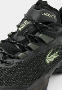 Černá sportovní obuv Lacoste s texturovaným svrškem, zelenými akcenty a reflexními tkaničkami. Má logo na bocích a polstrovaný límec.