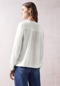 Blusa grigia chiaro a maniche lunghe in tessuto testurizzato, con scollatura rotonda e un taglio fluido. Indossata con jeans di denim blu.