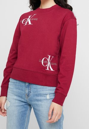 Kobieta w czerwonej bluzie Calvin Klein Jeans z logo na piersi i dole, zestawiona z jasnoniebieskimi, postarzanymi dżinsami.
