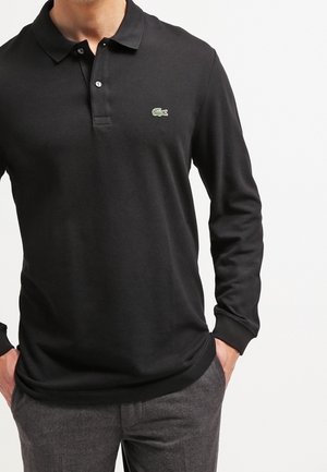 Polo noir à manches longues avec un col, une patte de boutonnage et un logo vert sur la poitrine gauche ; fabriqué en tissu lisse et léger.
