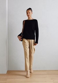 Schwarzer Pullover mit langen Ärmeln, beiger, plissierter Hose und nude-farbenen Stiefeletten. Das Model trägt eine braune Schultertasche vor einem hellen Hintergrund.