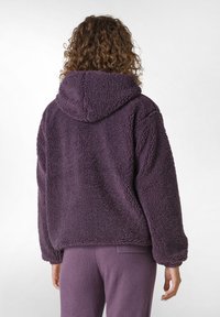 Hanorac din fleece violet, cu o suprafață texturată și pufoasă, având glugă. Mâneci lungi și o croială lejeră, asortat cu pantaloni corespunzători.