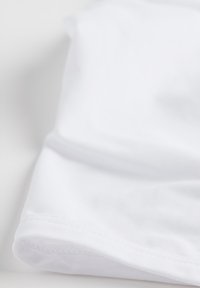 Biała tkanina o gładkiej fakturze, z widoczną złożoną krawędzią i delikatnym detalem przeszycia. Materiał wydaje się miękki i lekki.