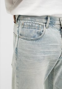 Hellblaue Jeans aus Denim, getragen mit einem weißen Hemd, Nahaufnahme der vorderen rechten Tasche und der Gürtelschlaufen.