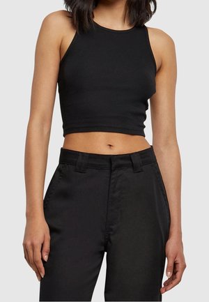 Vrouw in een zwart mouwloos crop top en zwarte broek met hoge taille, staand met ontspannen armen tegen een effen lichte achtergrond.