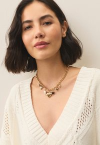 Gold necklace featuring multiple heart-shaped charms and a pearl accent on a textured chain, worn over a white knit sweater with a V-neck.