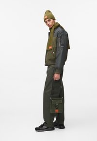 Ελαιοπράσινη μόνωτη ζακέτα με γκρι μανίκια, ταιριαστό σάλι, πράσινα παντελόνια, μαύρα αθλητικά και ελαιοπράσινη τσάντα με πορτοκαλί λεπτομέρεια.