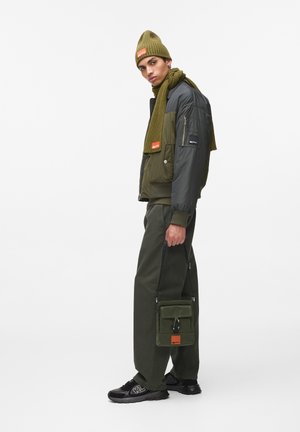 Olivgrüne, isolierte Jacke mit grauen Ärmeln, passendem Schal, grünen Hosen, schwarzen Sneakers und einer olivfarbenen Tasche mit orangefarbenen Akzenten.