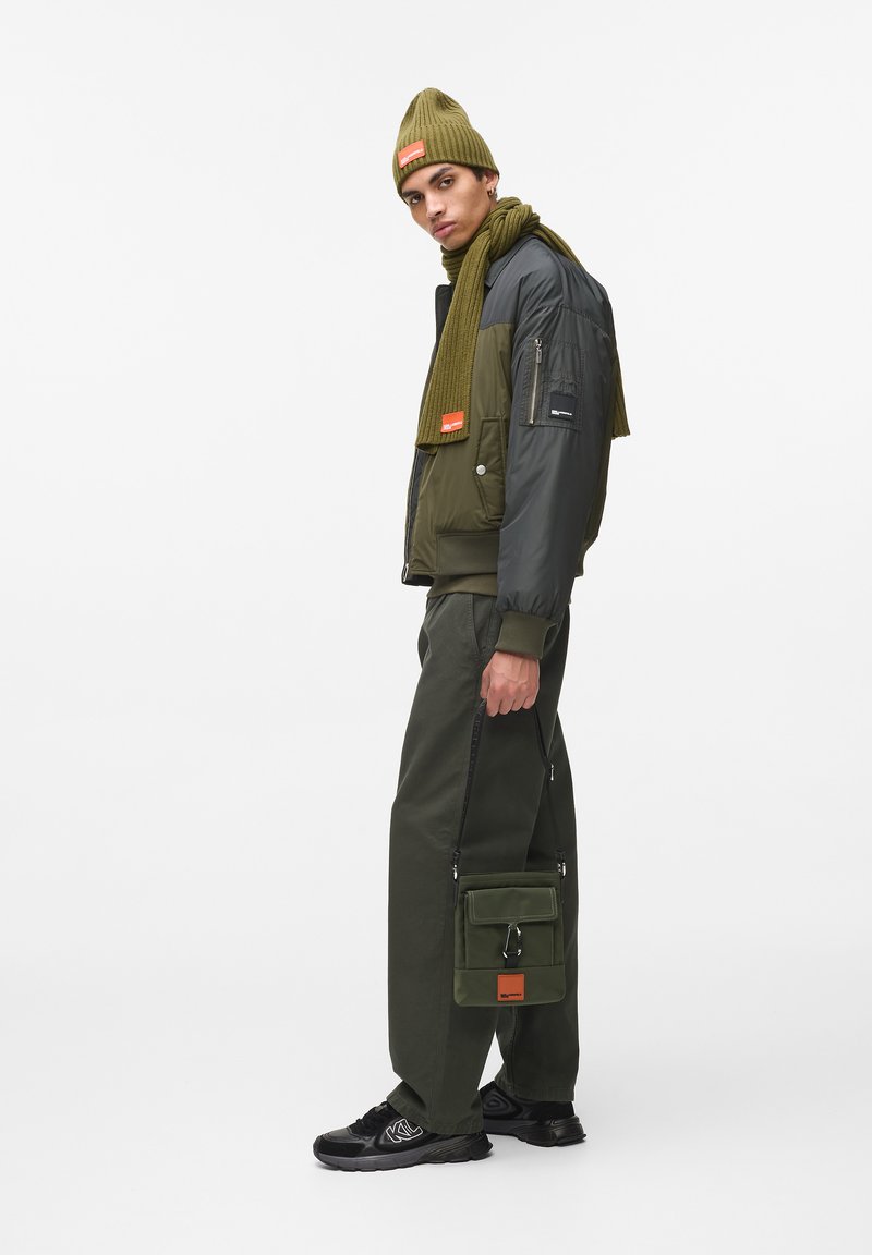 Ελαιοπράσινη μόνωτη ζακέτα με γκρι μανίκια, ταιριαστό σάλι, πράσινα παντελόνια, μαύρα αθλητικά και ελαιοπράσινη τσάντα με πορτοκαλί λεπτομέρεια.