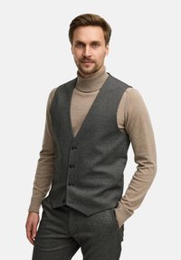 Grå mønstret vest med V-hals design og sorte knapper, båret over en beige turtleneck sweater. En skræddersyet pasform er tydelig.