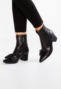 Botas de cuero negras hasta el tobillo con punta afilada y tacón cuadrado, con un lazo decorativo en la parte frontal y una cremallera lateral. Textura suave.