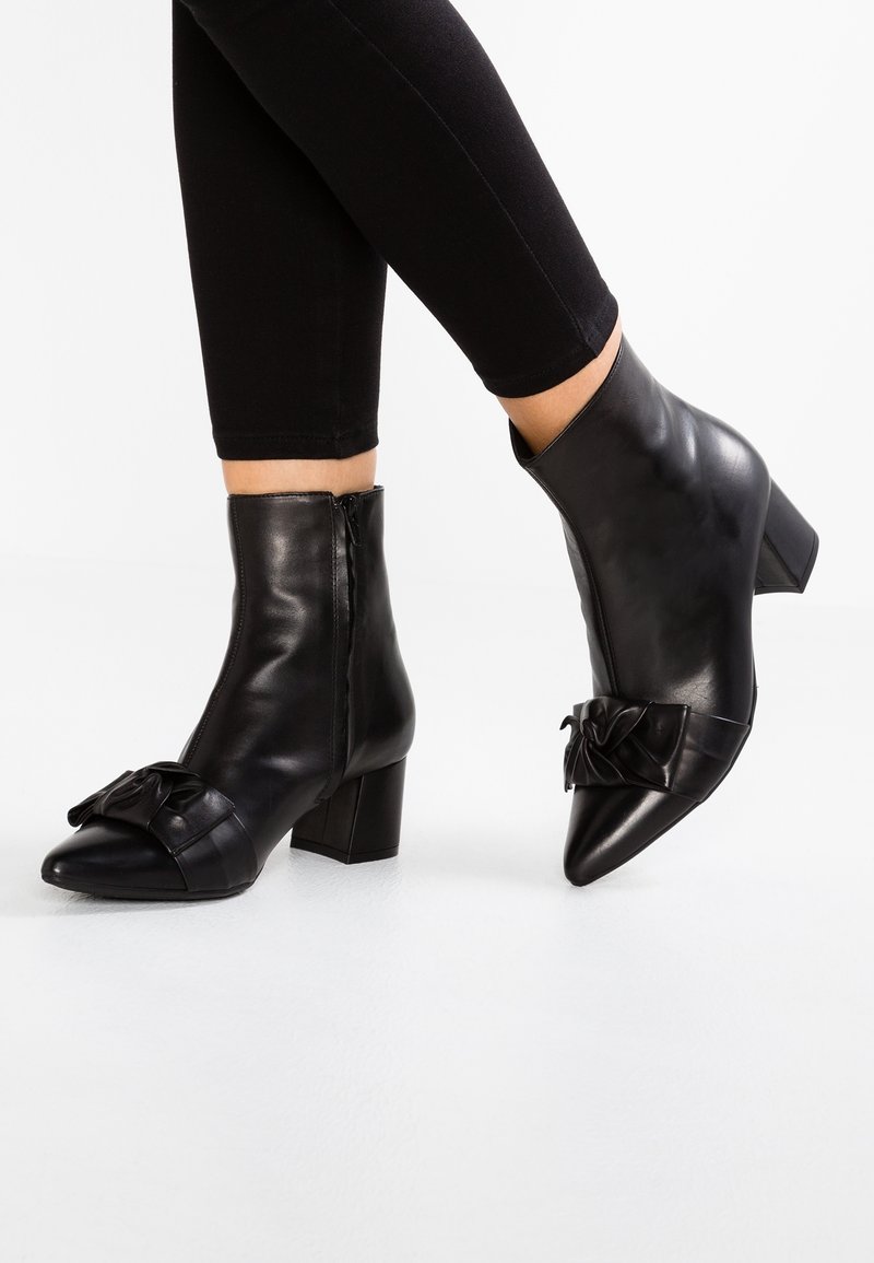 Botas de cuero negras hasta el tobillo con punta afilada y tacón cuadrado, con un lazo decorativo en la parte frontal y una cremallera lateral. Textura suave.