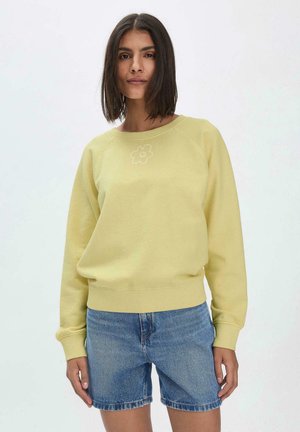 Hellgelbes Sweatshirt mit Rundhalsausschnitt und Raglanärmeln, ausgestattet mit einem gestickten Blumenmotiv auf der Brust. Kombiniert mit Denim-Shorts.