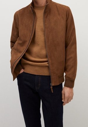 Homme portant une veste zippée en daim marron sur un pull marron clair et un jean bleu foncé, debout avec une main dans la poche.