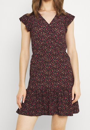 Femme portant une robe noire à fleurs, manches courtes, col en V et ourlet volanté, debout devant un fond blanc uni.