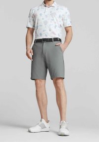 Bílá polokošile s barevnými vzory palem a nápojů, doplněná o šedé šortky. Černý pásek a bílé golfové boty se šedými detaily.