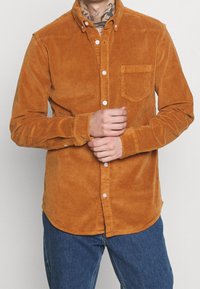Chemise en velours côtelé couleur orange brûlé, dotée d'une poche poitrine, d'un col pointu et de boutons blancs, associée à un jean bleu.