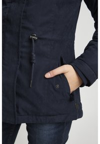 Navy blue pullover jacket made of textured fabric, featuring a drawstring at the waist, side pockets, and a snap button accent.
