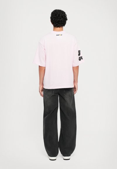 Ružová tričko s krátkym rukávom a čiernym nápisom 'NAPOLI' na chrbte, doplnené grafikou na rukáve, skombinované so širokými tmavosivými džínsami a bielymi teniskami.