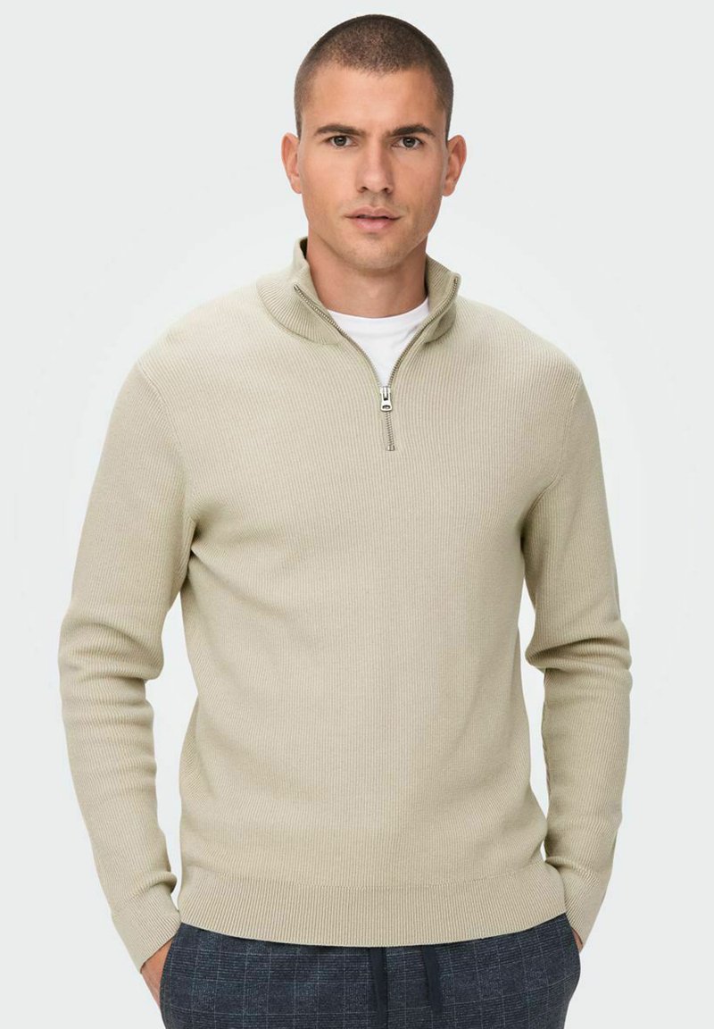 Suéter acanalado de color beige claro con cuello de media cremallera, tejido texturizado y mangas ajustadas, combinado con pantalones de cuadros grises.