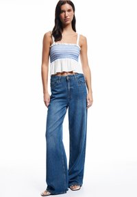 Witte crop top met blauwe borduursels, gecombineerd met brede blauwe spijkerbroeken. De outfit heeft een gestructureerde stof en een vloeiende uitstraling.