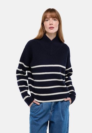 Pull côtelé bleu marine avec des rayures horizontales blanches, doté d'un col montant et d'une coupe décontractée. Associé à un jean bleu à jambes larges.
