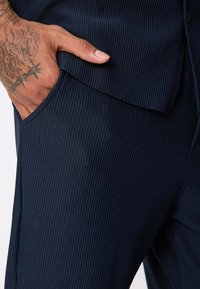 Pantalons en tissu côtelé bleu marine avec un détail de poche et une main reposant sur le côté. Texture visible et design ajusté.