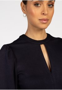 Frau mit Kreole, die ein schwarzes Top mit einem kleinen Ausschnitt vorne trägt, sichtbares Makeup und neutralen Lipgloss hat.