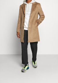 Man som bär en beige överrock, vit huvtröja med snoddar, svarta byxor och sneakers med neonaccenter, står framför en enkel bakgrund.