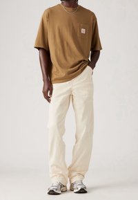 T-shirt marrone a maniche corte con tasca frontale, abbinato a pantaloni beig larghi e sneaker argento con accenti neri.